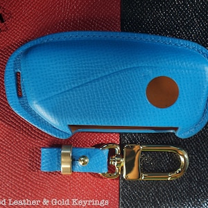 May include: A blue leather key fob with a gold keyring. The key fob has a textured surface and a small circle cutout. "*Textured Leather & Gold Keyrings" is written at the bottom of the image.