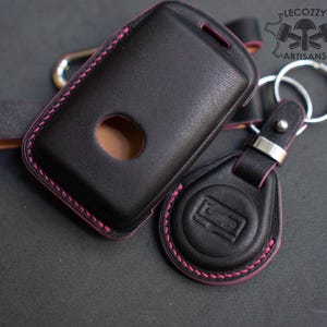 Può includere: Portachiavi e custodia in pelle nera con cuciture rosa. Il portachiavi ha un anello argentato e un cinturino in pelle nera. La custodia ha un ritaglio e un logo. Il logo dice "LECOZZY ARTISANS".