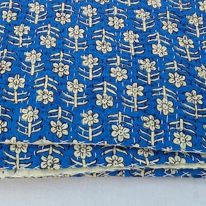 May include: Folded blue fabric featuring a cream and navy floral pattern. The textile, possibly a quilt, shows stitching details. The design is symmetrical and decorative. The fabric is likely intended for crafting or home decor.
