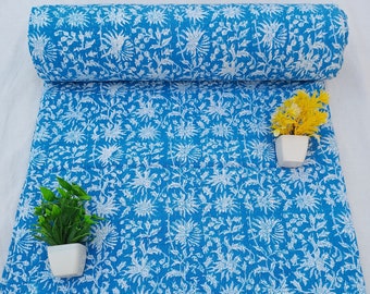 May include: A blue and white kantha quilt fabric with a floral pattern. The fabric is rolled up and there are two small potted plants in the image.