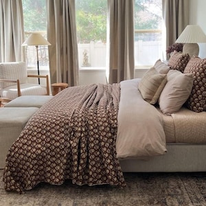 Puede incluir: Interior de dormitorio con una cama vestida con un edred&oacute;n floral marr&oacute;n y cojines. La cama tiene una funda n&oacute;rdica rosa claro y varias almohadas decorativas. Una alfombra estampada y cortinas neutras crean un ambiente c&aacute;lido y acogedor.