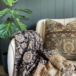May include: A black and cream patterned throw blanket with fringe edges draped over a white armchair with a patterned pillow. The armchair is in front of a green wood paneled wall.