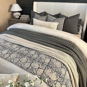 May include: A bed featuring a light-colored headboard, a white duvet, a black and white floral patterned runner, and a thick dark gray knitted throw blanket. The bed is layered with several pillows in shades of gray, white, and black.