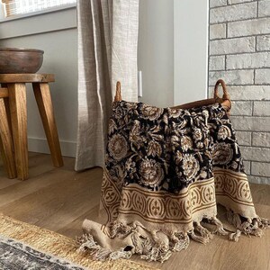 May include: A black and brown throw blanket with a floral pattern and tassels. The blanket is folded and placed in a wicker basket.