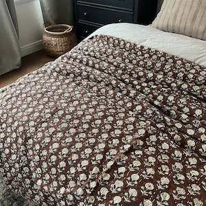 Puede incluir: Un edred&oacute;n marr&oacute;n con estampado floral sobre una cama. El edred&oacute;n presenta un patr&oacute;n repetido de flores blancas y est&aacute; cosido con hilo blanco. La cama est&aacute; hecha con una s&aacute;bana blanca y una almohada a rayas. Una c&oacute;moda negra y una cesta de mimbre est&aacute;n en el fondo.