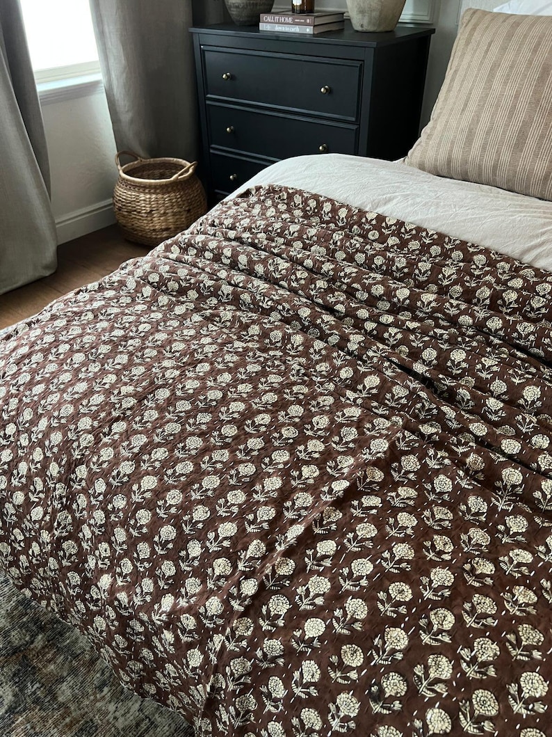 Puede incluir: Un edred&oacute;n marr&oacute;n con estampado floral sobre una cama. El edred&oacute;n presenta un patr&oacute;n repetido de flores blancas y est&aacute; cosido con hilo blanco. La cama est&aacute; hecha con una s&aacute;bana blanca y una almohada a rayas. Una c&oacute;moda negra y una cesta de mimbre est&aacute;n en el fondo.