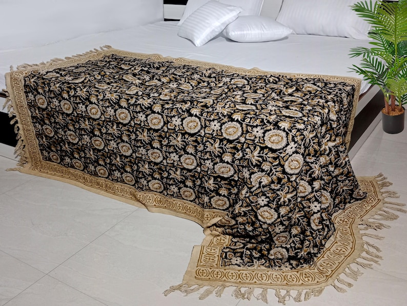 May include: Black and cream floral patterned throw blanket with a fringe border. The blanket is draped over a bed with white pillows.