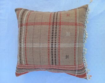 May include: A brown and red plaid cushion with white fringe trim. The cushion has a woven pattern with geometric designs.