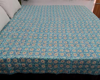 Puede incluir: Una colcha turquesa con un estampado floral repetido en tonos azules y dorados. La colcha está extendida sobre una cama con almohadas blancas. El diseño floral es una serie de formas circulares.