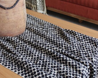 May include: A black and white patterned throw blanket with a fringe edge is draped over a wooden coffee table. A large terracotta vase with a rope handle sits on the table.