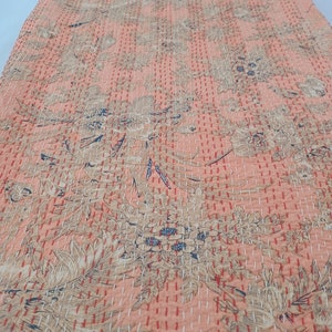 May include: A peach-colored textile with a floral pattern and intricate stitching. The design features large, stylized flowers and leaves in shades of beige and blue. The stitching is in a contrasting white and red thread, creating a textured effect.