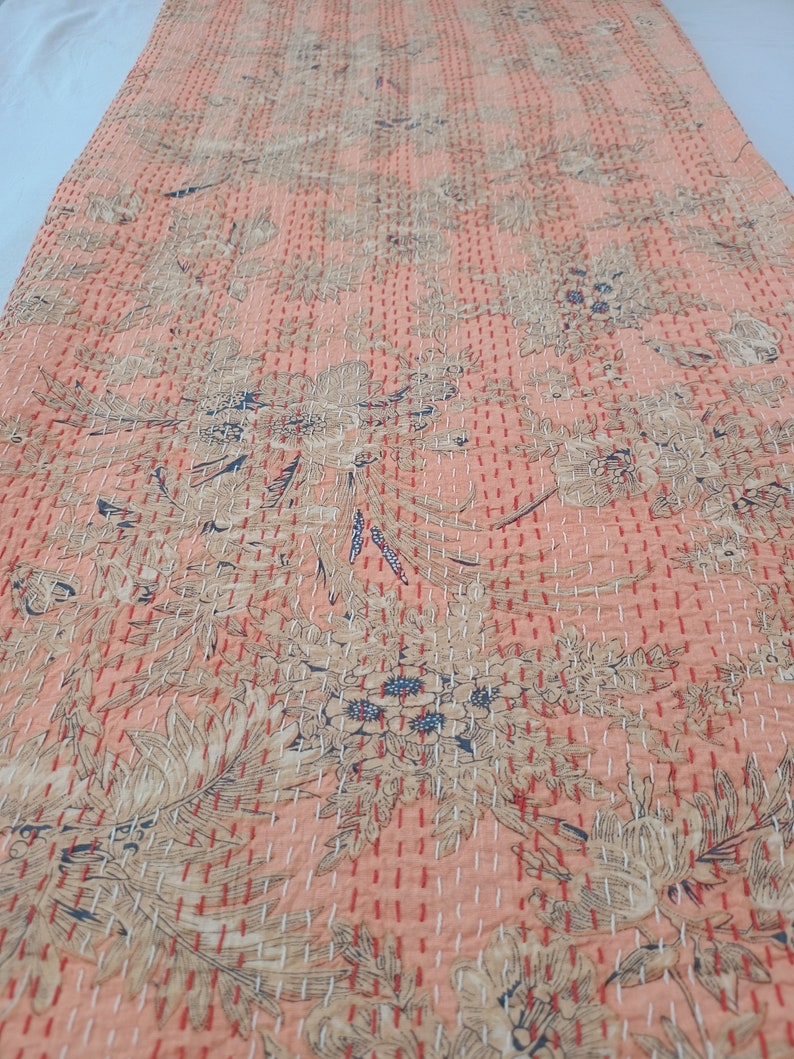 May include: A peach-colored textile with a floral pattern and intricate stitching. The design features large, stylized flowers and leaves in shades of beige and blue. The stitching is in a contrasting white and red thread, creating a textured effect.