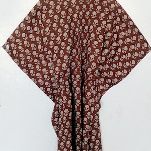 Puede incluir: Un vestido caft&aacute;n marr&oacute;n con estampado floral, colgado de una percha de madera. La tela presenta un dise&ntilde;o repetido de peque&ntilde;as flores blanquecinas sobre un fondo marr&oacute;n oscuro. El vestido se muestra contra una pared blanca.