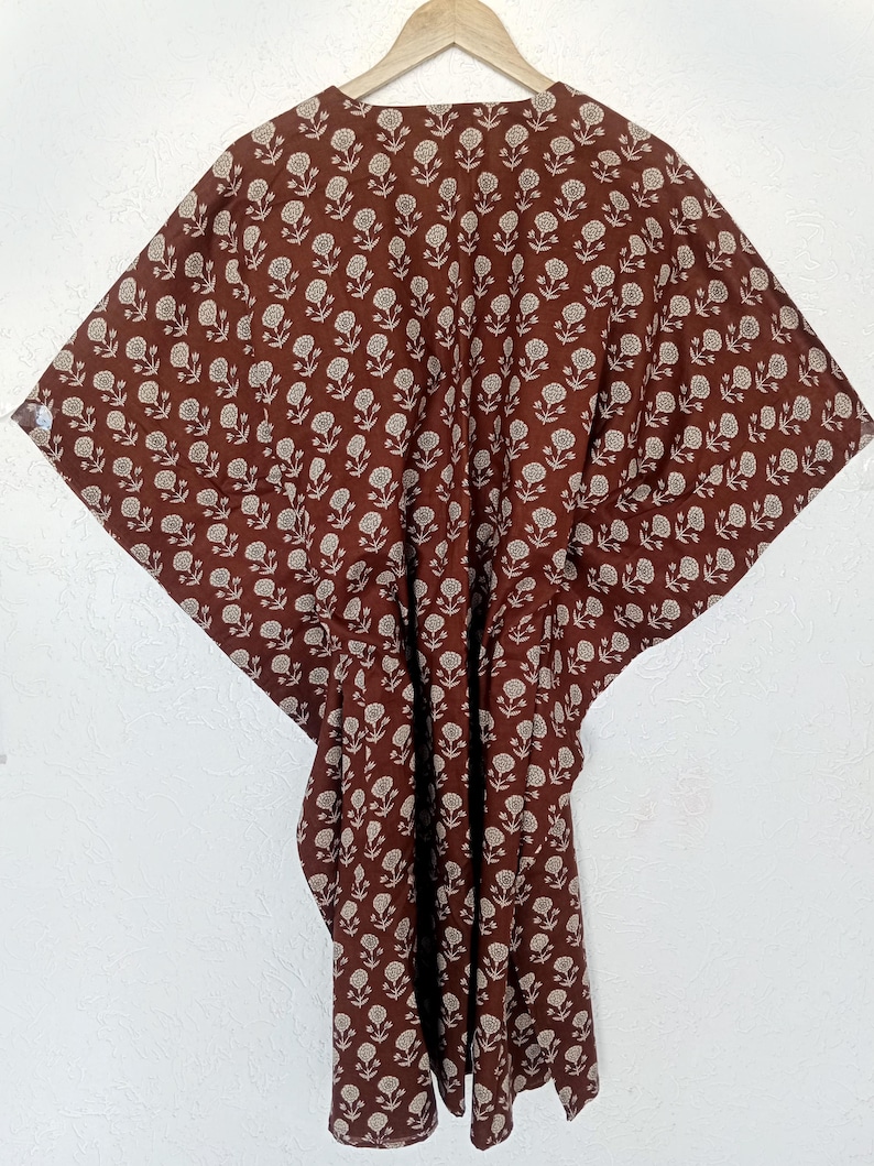 Puede incluir: Un vestido caft&aacute;n marr&oacute;n con estampado floral, colgado de una percha de madera. La tela presenta un dise&ntilde;o repetido de peque&ntilde;as flores blanquecinas sobre un fondo marr&oacute;n oscuro. El vestido se muestra contra una pared blanca.