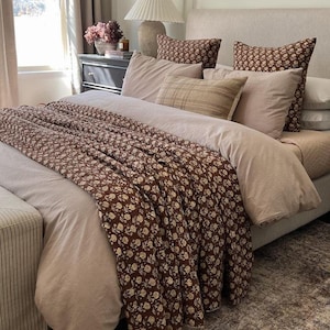 Puede incluir: Un dormitorio con una cama con ropa de cama de tonos neutros y una manta con estampado floral marr&oacute;n. Cojines a juego adornan la cama. Una alfombra estampada y una mesita de noche con una l&aacute;mpara completan la decoraci&oacute;n.