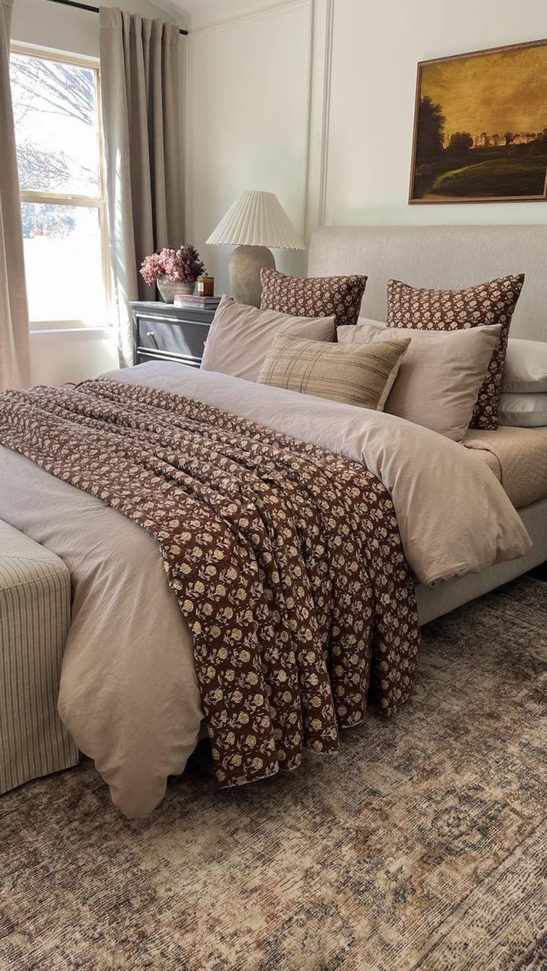 Puede incluir: Un dormitorio con una cama con ropa de cama de tonos neutros y una manta con estampado floral marr&oacute;n. Cojines a juego adornan la cama. Una alfombra estampada y una mesita de noche con una l&aacute;mpara completan la decoraci&oacute;n.