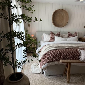 Puede incluir: Interior de dormitorio con una cama vestida con un edred&oacute;n floral burdeos y blanco, almohadas blancas y una manta crema. Un banco de madera est&aacute; al pie de la cama. Una gran planta en maceta y un tapiz de pared contribuyen a la decoraci&oacute;n de la habitaci&oacute;n.