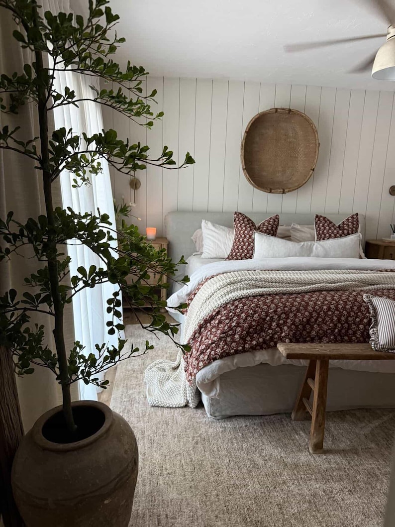 Puede incluir: Interior de dormitorio con una cama vestida con un edred&oacute;n floral burdeos y blanco, almohadas blancas y una manta crema. Un banco de madera est&aacute; al pie de la cama. Una gran planta en maceta y un tapiz de pared contribuyen a la decoraci&oacute;n de la habitaci&oacute;n.