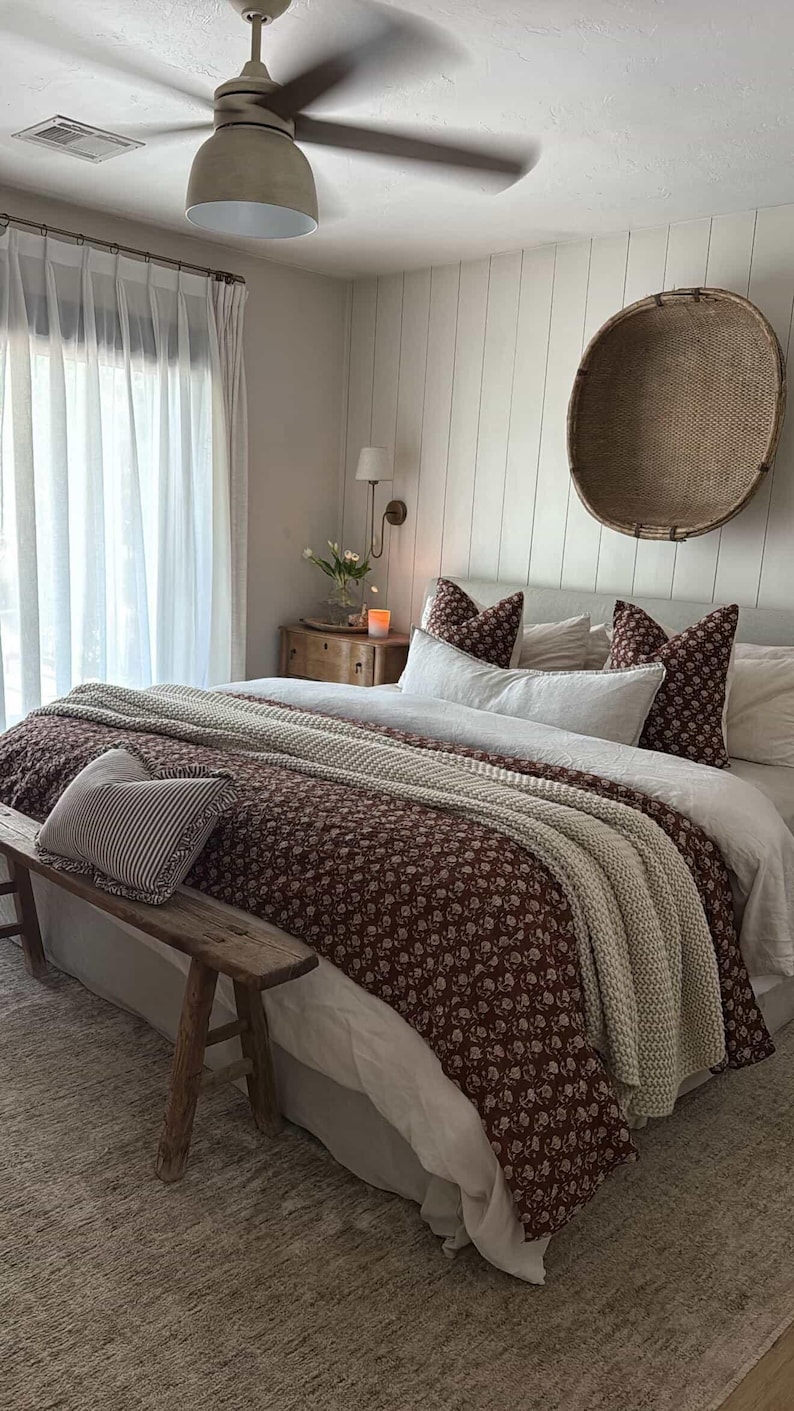 Puede incluir: Interior de dormitorio con una cama vestida con un edred&oacute;n floral y cojines. Una manta de punto de color crema y un coj&iacute;n a rayas a&ntilde;aden textura. Un banco de madera est&aacute; al pie de la cama y una cesta tejida cuelga de la pared.