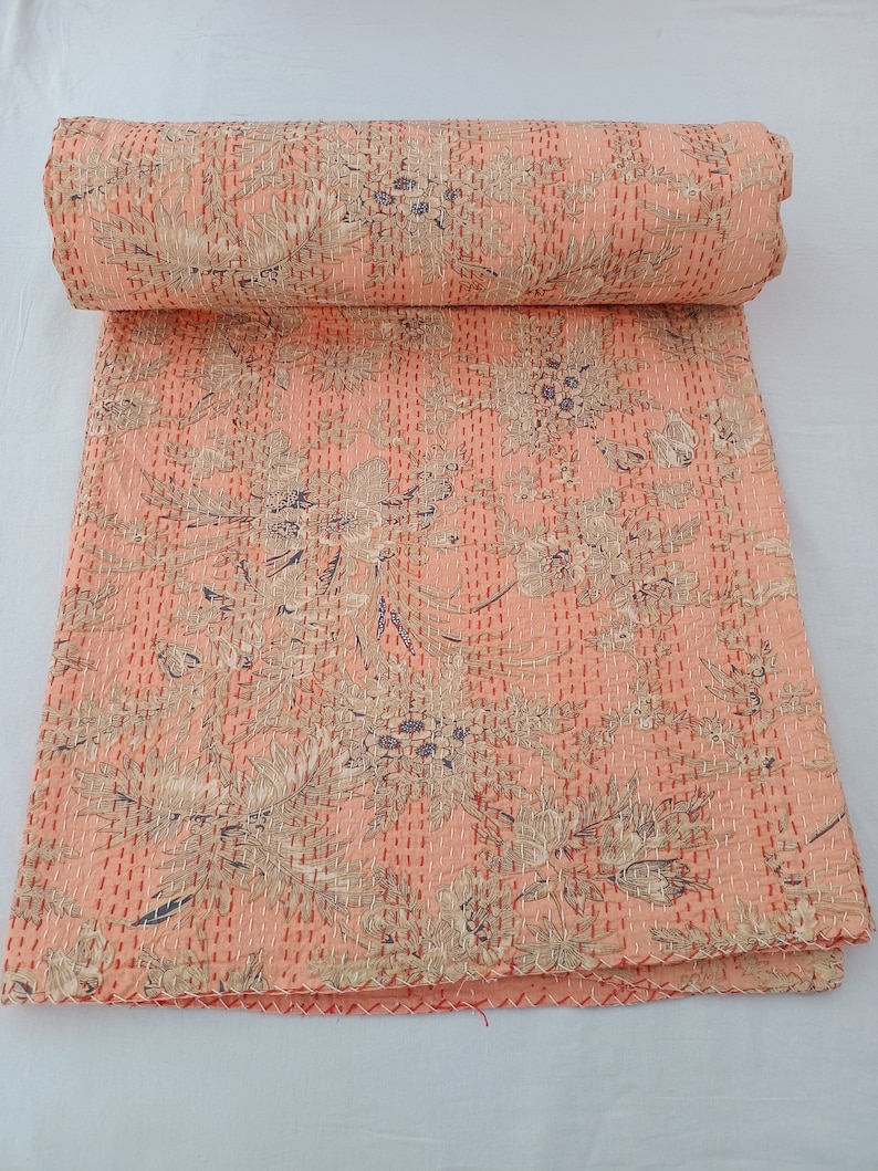 May include: A rolled-up peach-colored quilt with a floral pattern in beige and blue. The quilt is hand-stitched with white thread, and the edges are finished with a red stitch.