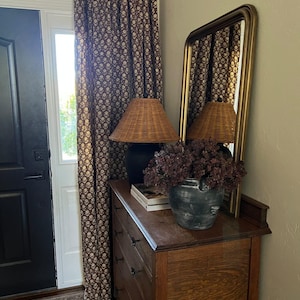 Puede incluir: Interior de una habitaci&oacute;n con una c&oacute;moda de madera, un espejo con marco dorado y una l&aacute;mpara de mimbre. Un jarr&oacute;n negro con flores secas descansa sobre la c&oacute;moda. Cortinas marrones con estampado floral cuelgan junto a una puerta blanca.