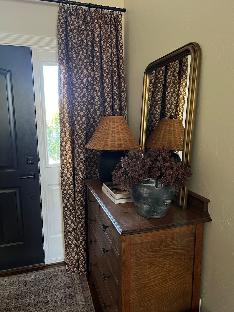 Puede incluir: Interior de una habitaci&oacute;n con una c&oacute;moda de madera, un espejo con marco dorado y una l&aacute;mpara de mimbre. Un jarr&oacute;n negro con flores secas descansa sobre la c&oacute;moda. Cortinas marrones con estampado floral cuelgan junto a una puerta blanca.