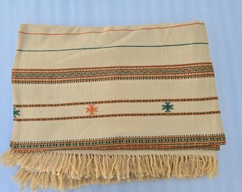 Puede incluir: Textil tejido de color crema con rayas horizontales en naranja, verde y marrón. El textil presenta patrones geométricos bordados y flecos a lo largo del borde inferior. El textil doblado se muestra sobre una superficie azul claro.