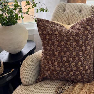 Puede incluir: Un coj&iacute;n decorativo con un estampado floral en tonos marr&oacute;n y beige descansa sobre un sill&oacute;n a rayas. El coj&iacute;n es de forma cuadrada y parece estar hecho de una tela suave. Una manta estampada est&aacute; sobre la silla.