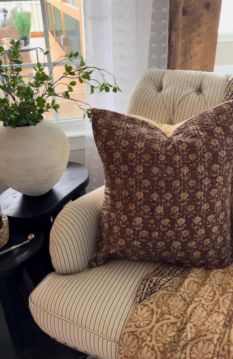 Puede incluir: Un coj&iacute;n decorativo con un estampado floral en tonos marr&oacute;n y beige descansa sobre un sill&oacute;n a rayas. El coj&iacute;n es de forma cuadrada y parece estar hecho de una tela suave. Una manta estampada est&aacute; sobre la silla.