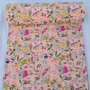 May include: A rolled-up, floral-patterned quilt in shades of peach, green, pink, and purple. The quilt features a repeating pattern of flowers and leaves, with white stitching throughout. The quilt is made of a soft, lightweight fabric.