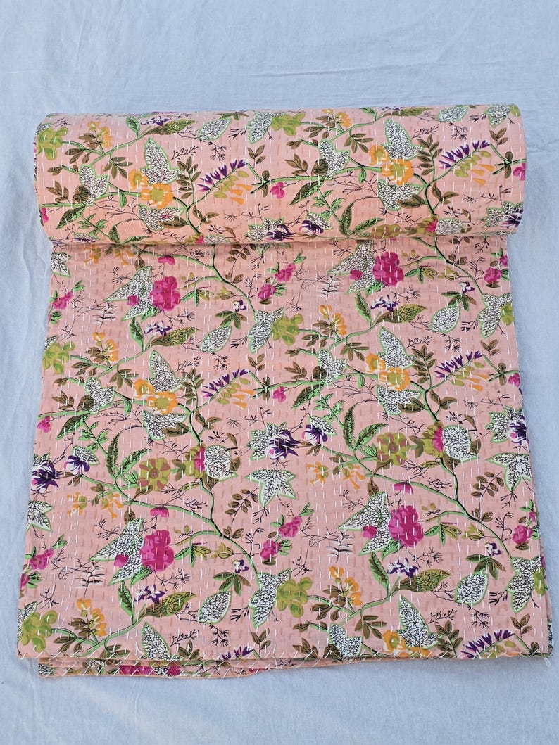 May include: A rolled-up, floral-patterned quilt in shades of peach, green, pink, and purple. The quilt features a repeating pattern of flowers and leaves, with white stitching throughout. The quilt is made of a soft, lightweight fabric.