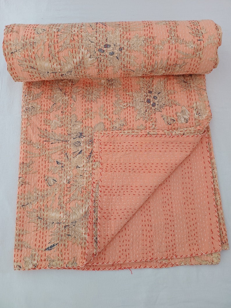May include: A peach-colored quilt with a floral pattern in beige and navy blue. The quilt is hand-stitched with red thread, and the edges are neatly finished. The quilt is folded and rolled, showcasing the intricate design and craftsmanship.