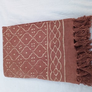 May include: A rust-colored cotton throw blanket with a white geometric pattern and fringe on the edges.