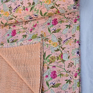 May include: A floral patterned quilt in shades of pink, green, purple, and yellow. The quilt features a repeating pattern of flowers and leaves, with white stitching throughout. The quilt is folded, revealing a solid peach-colored underside.