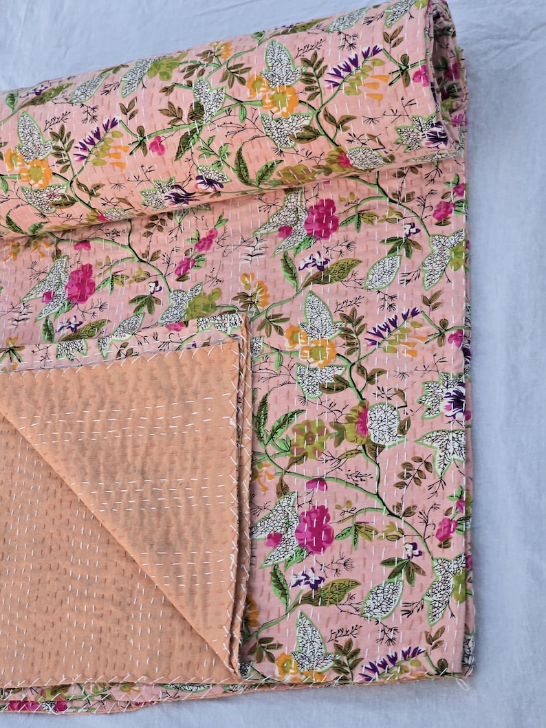May include: A floral patterned quilt in shades of pink, green, purple, and yellow. The quilt features a repeating pattern of flowers and leaves, with white stitching throughout. The quilt is folded, revealing a solid peach-colored underside.