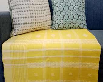 Könnte beinhalten: Eine gelbe Decke mit weißem Gittermuster und Fransen, drapiert über einem blauen Sofa. Zwei dekorative Kissen in Grau und Türkis liegen auf dem Sofa. Das graue Kissen hat ein gepunktetes Design, und das türkisfarbene Kissen hat ein geometrisches Muster.
