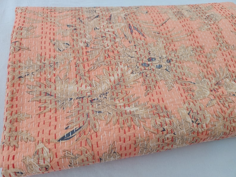 May include: A peach-colored quilt with a floral pattern in beige, red, and navy blue. The quilt features a repeating design of stylized flowers and foliage, with visible white stitching throughout. The overall aesthetic is vintage and handcrafted.