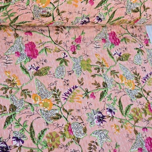 May include: A pink floral quilt with a repeating pattern of flowers and leaves in shades of pink, purple, green, and yellow. The quilt is hand-stitched with white thread, creating a textured surface. The overall design is a botanical theme.