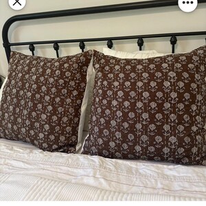 Puede incluir: Dos cojines decorativos marrones con un estampado floral blanco sobre una cama con ropa de cama blanca. Los cojines tienen una textura acolchada y est&aacute;n colocados contra un cabecero de metal negro. El tema general es la decoraci&oacute;n del hogar.