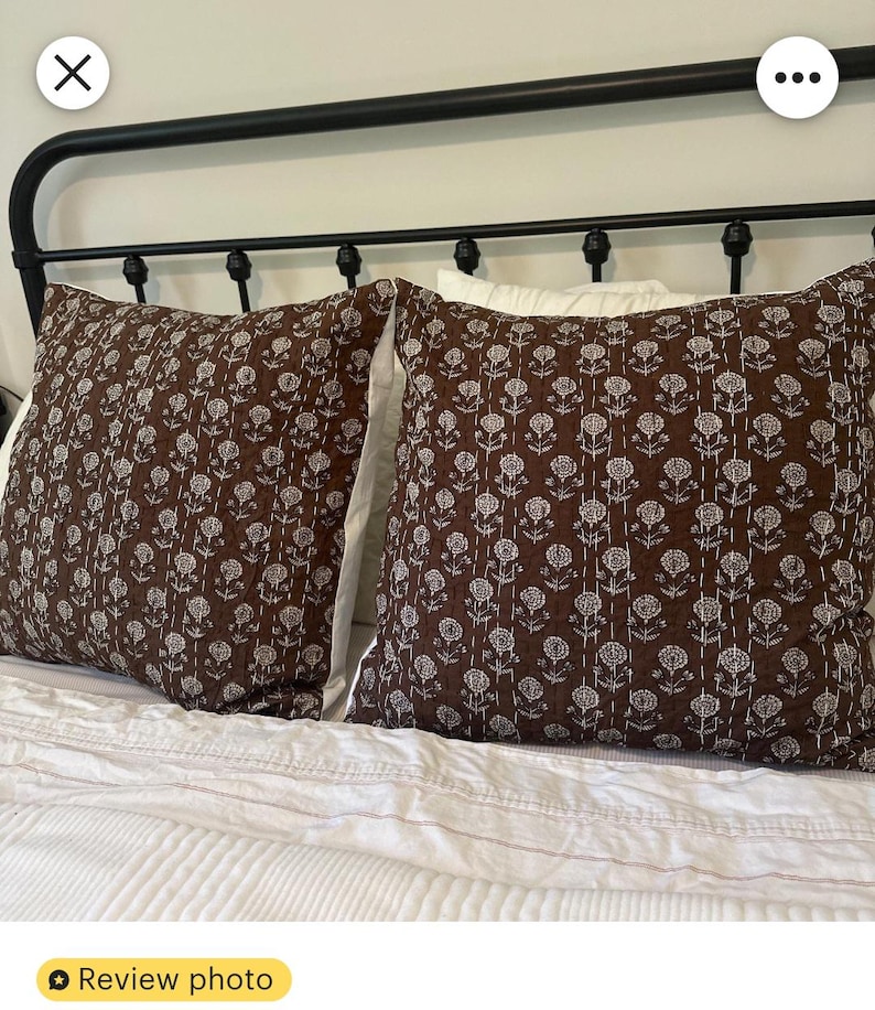 Puede incluir: Dos cojines decorativos marrones con un estampado floral blanco sobre una cama con ropa de cama blanca. Los cojines tienen una textura acolchada y est&aacute;n colocados contra un cabecero de metal negro. El tema general es la decoraci&oacute;n del hogar.