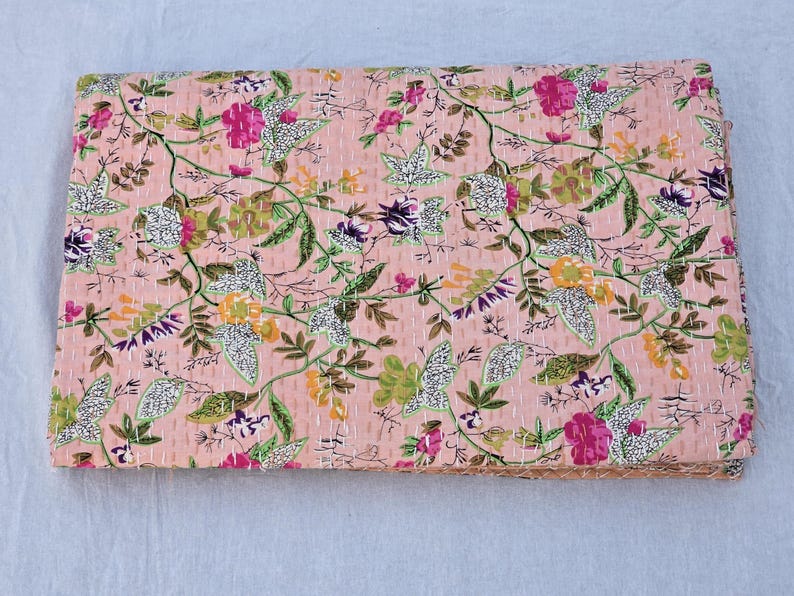 May include: A folded, floral-patterned quilt in shades of pink, green, yellow, and purple. The quilt features a repeating pattern of flowers and leaves, with visible stitching throughout. The edges are neatly finished.