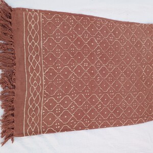 May include: A terracotta-colored cotton throw blanket with a white geometric pattern and fringe trim.