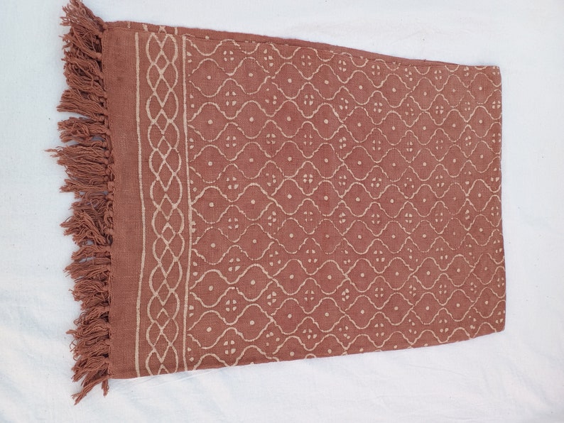 May include: A terracotta-colored cotton throw blanket with a white geometric pattern and fringe trim.