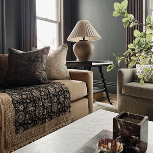 May include: A living room with a brown couch, a black and gold throw blanket, a white lamp with a cream shade, and a marble coffee table with a book and a decorative candle holder.