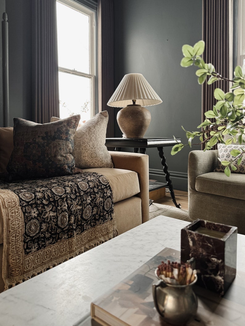 May include: A living room with a brown couch, a black and gold throw blanket, a white lamp with a cream shade, and a marble coffee table with a book and a decorative candle holder.