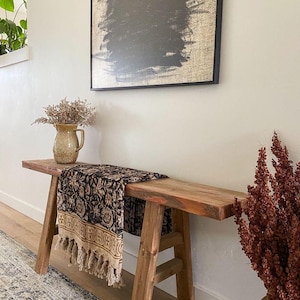 May include: A rustic wooden bench with a black and brown patterned throw blanket draped over it. The bench is in a hallway with a vase of dried flowers and a framed abstract painting on the wall.