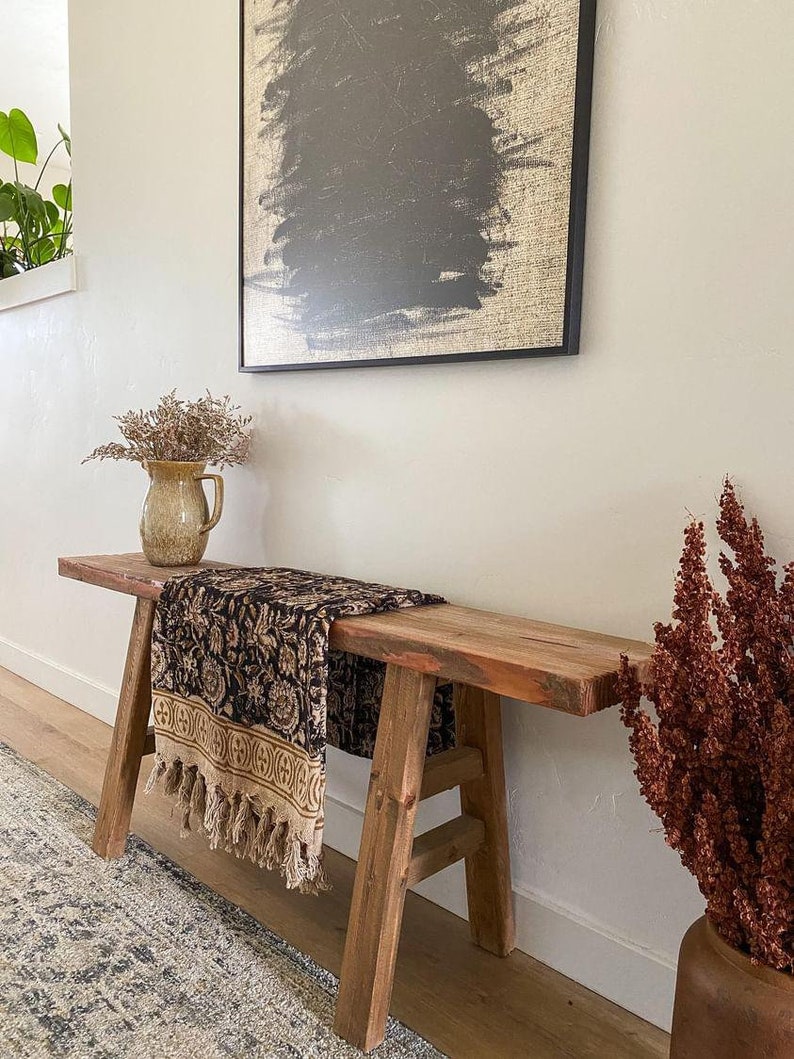 May include: A rustic wooden bench with a black and brown patterned throw blanket draped over it. The bench is in a hallway with a vase of dried flowers and a framed abstract painting on the wall.