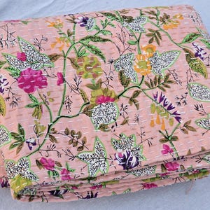 May include: A folded, pink floral quilt with a repeating pattern of flowers and leaves in shades of pink, green, purple, and yellow. The quilt is stitched with white thread, creating a textured effect. The overall design is a botanical theme.
