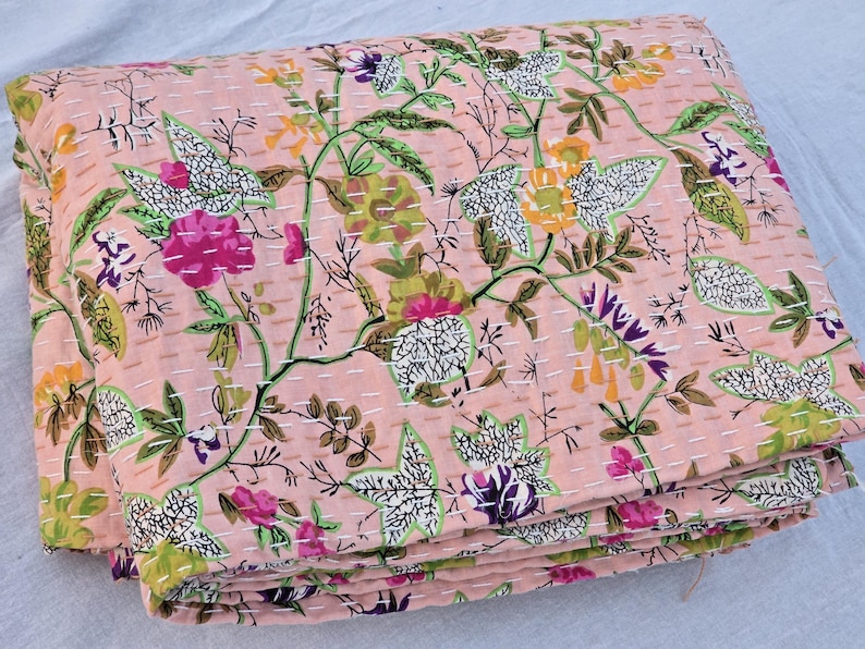 May include: A folded, pink floral quilt with a repeating pattern of flowers and leaves in shades of pink, green, purple, and yellow. The quilt is stitched with white thread, creating a textured effect. The overall design is a botanical theme.