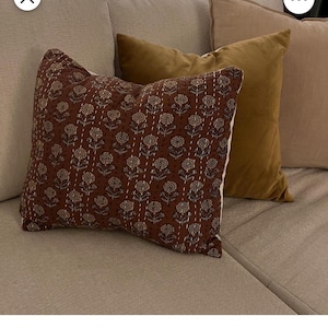 Puede incluir: Un coj&iacute;n decorativo con un estampado floral marr&oacute;n y costuras blancas. El coj&iacute;n est&aacute; sobre un sof&aacute; beige, con otros dos cojines dorados y beige. Los cojines a&ntilde;aden un toque de elegancia al sal&oacute;n.